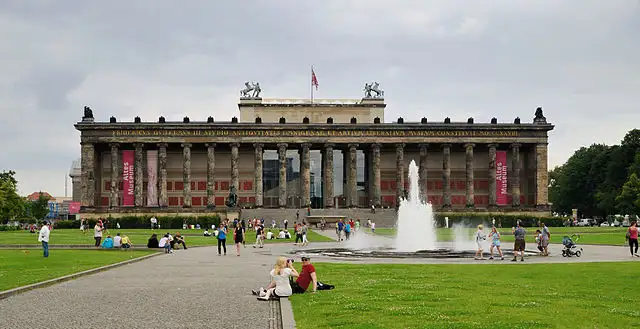 The Altes Museum on an overcast day. The long building is lined with columns. In front of the museum, people relax around a fountain and on the green grass.