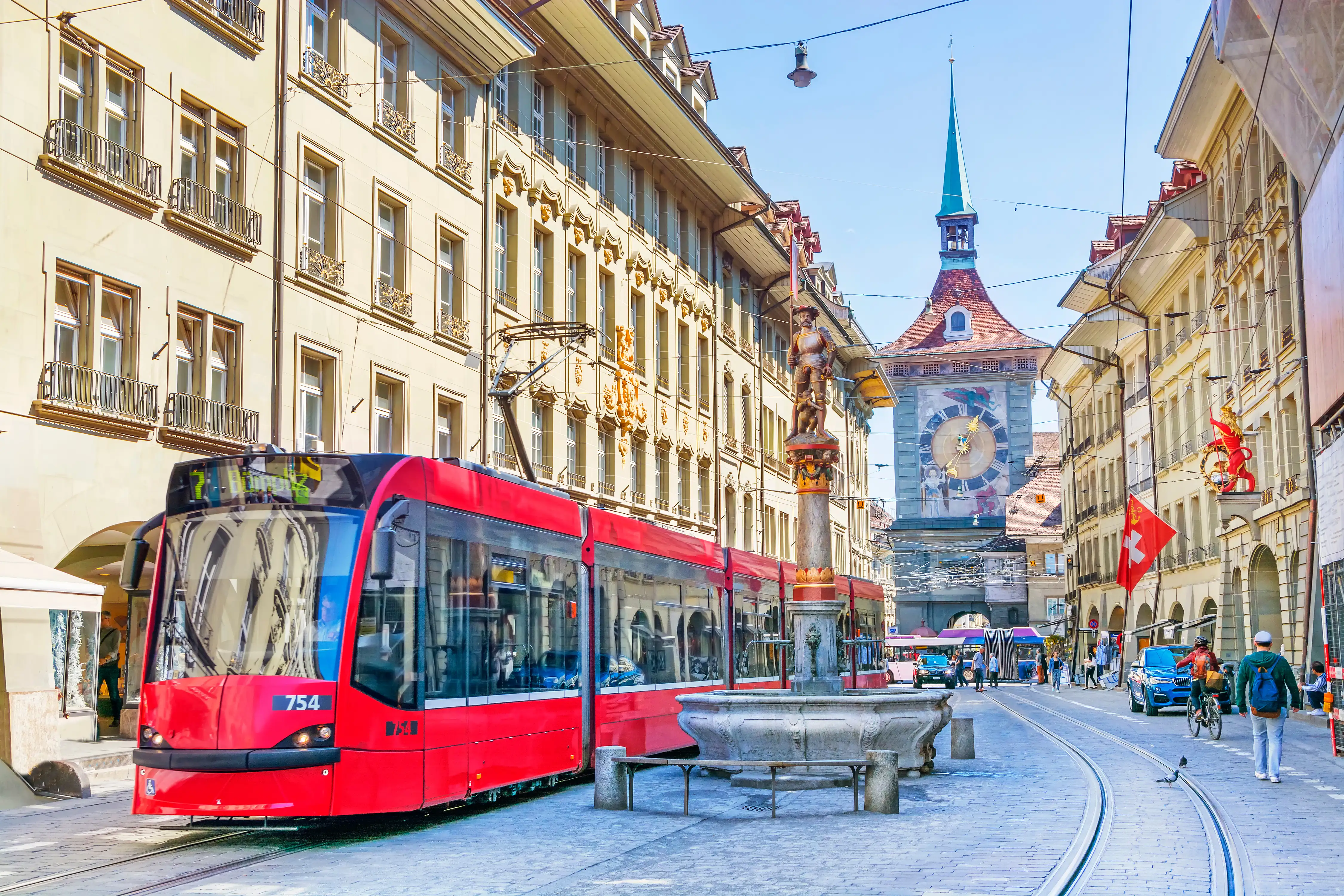 A large historic clock tower in an old European city centre with a red tram travelling through the street