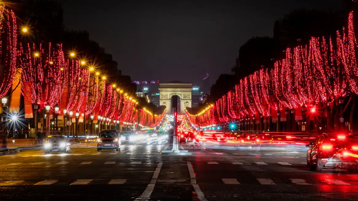 Nighttime view of the Champs-Élysées Christmas lights, with red-lit trees lining the road and blurred car lights in the distance.