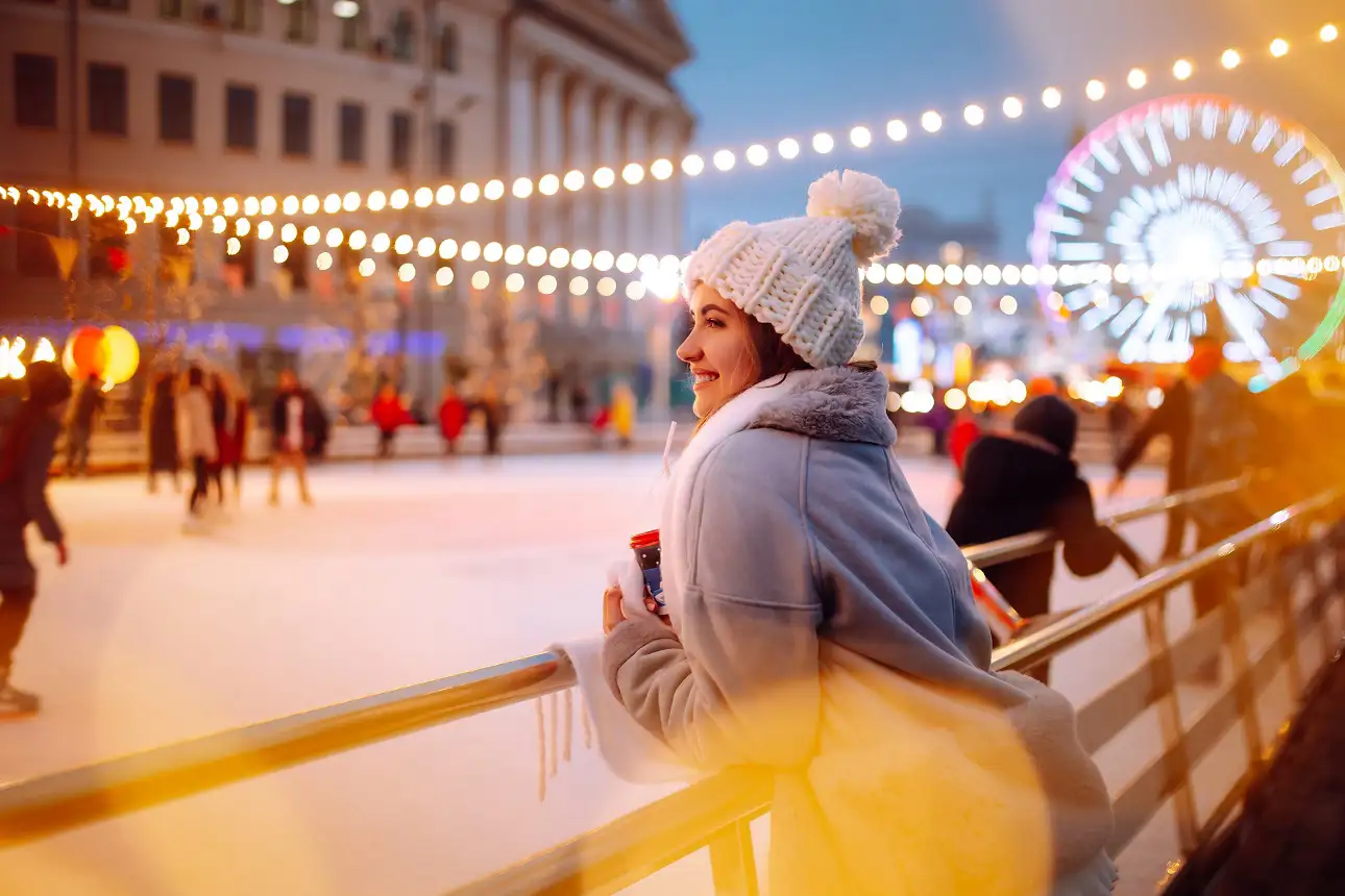 Smiling woman in winter clothing holding coffee near a skating rink, surrounded by glowing Christmas lights.