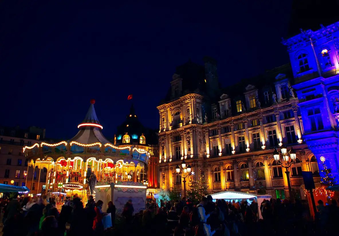 Night view of the Hôtel de Ville Christmas market, featuring a brightly lit carousel, the illuminated Hôtel de Ville building, and people enjoying the festive atmosphere.