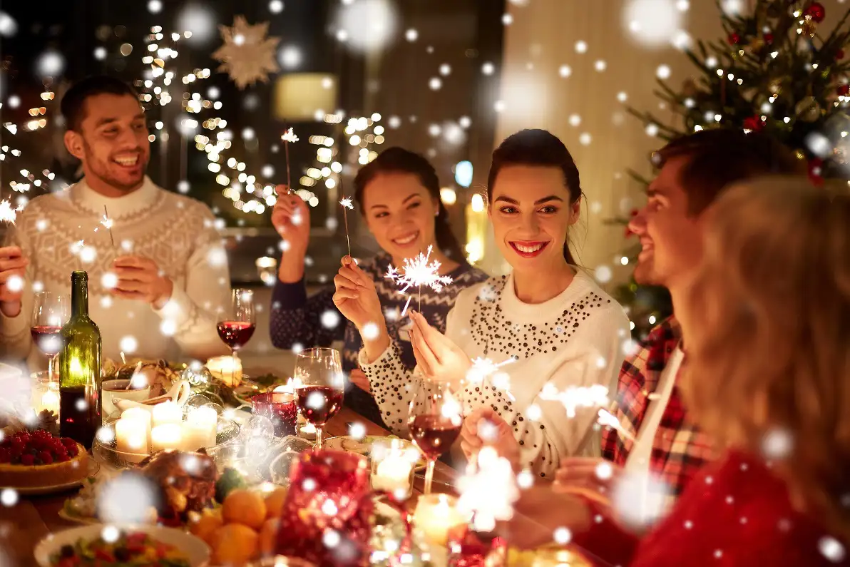 Group of friends sitting at a Christmas dinner table, holding sparklers, surrounded by pretty lights and holiday decorations.