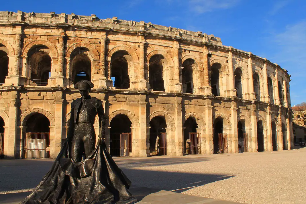 Les Arènes in Arles under a clear blue sky. In the foreground is a sculpture of a matador