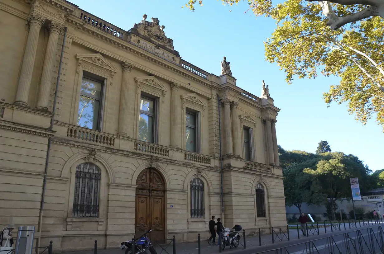 View of Musée Fabre during the day from a parallel perspective, featuring a clear blue sky, vibrant foliage, and pedestrians walking by