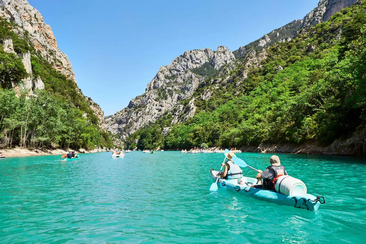 Gorges du Verdon with clear turquoise water viewed from the water's perspective, under blue skies, surrounded by lush green foliage and rocky cliffs. People in canoes float on the water.