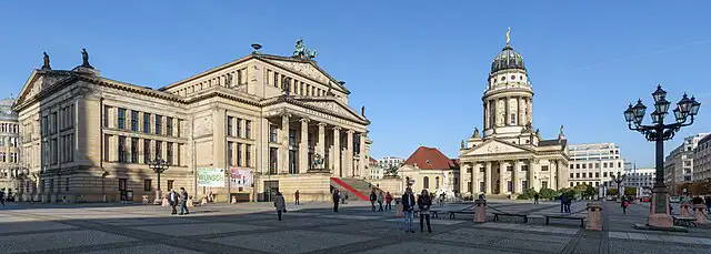 A large-paved square with a view of some of monumental buildings that surround it. The historic concert hall and French cathedrals are visible against a blue sky. People are walking around the space.