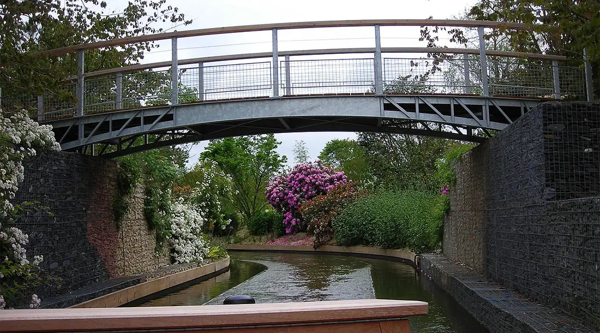 A bridge over a narrow man made river or canal, with colourful flowers and shrubs on both banks.