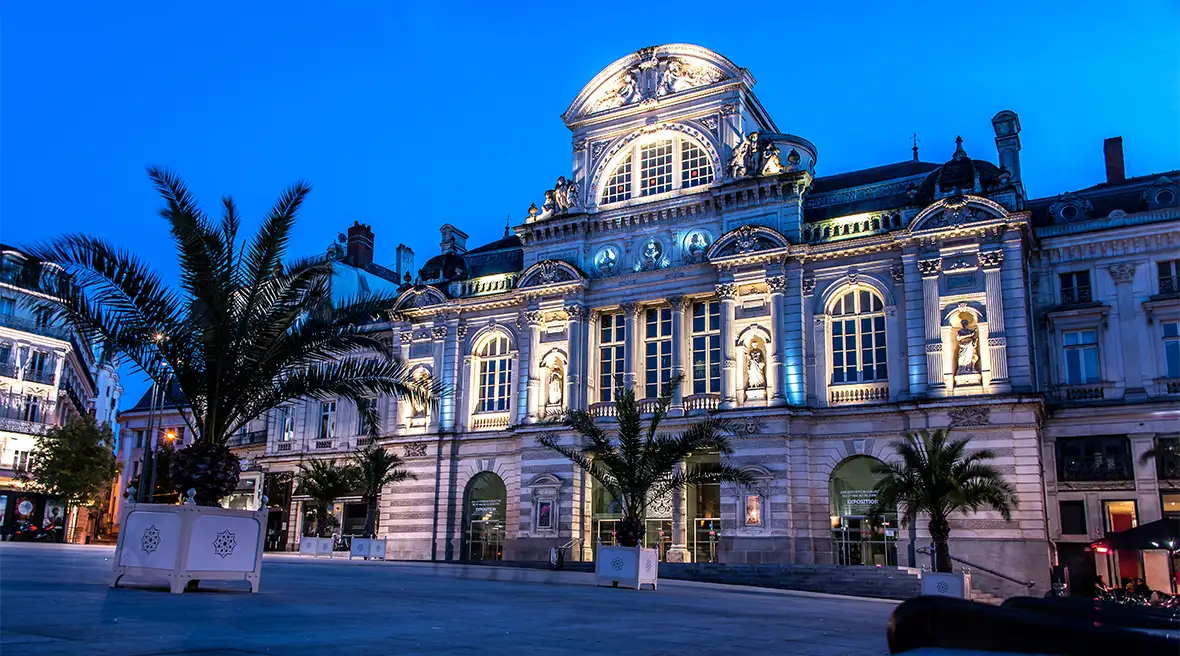 A softly illuminated grand building with a palatial frontage on a large square at night, in front of it are tropical trees