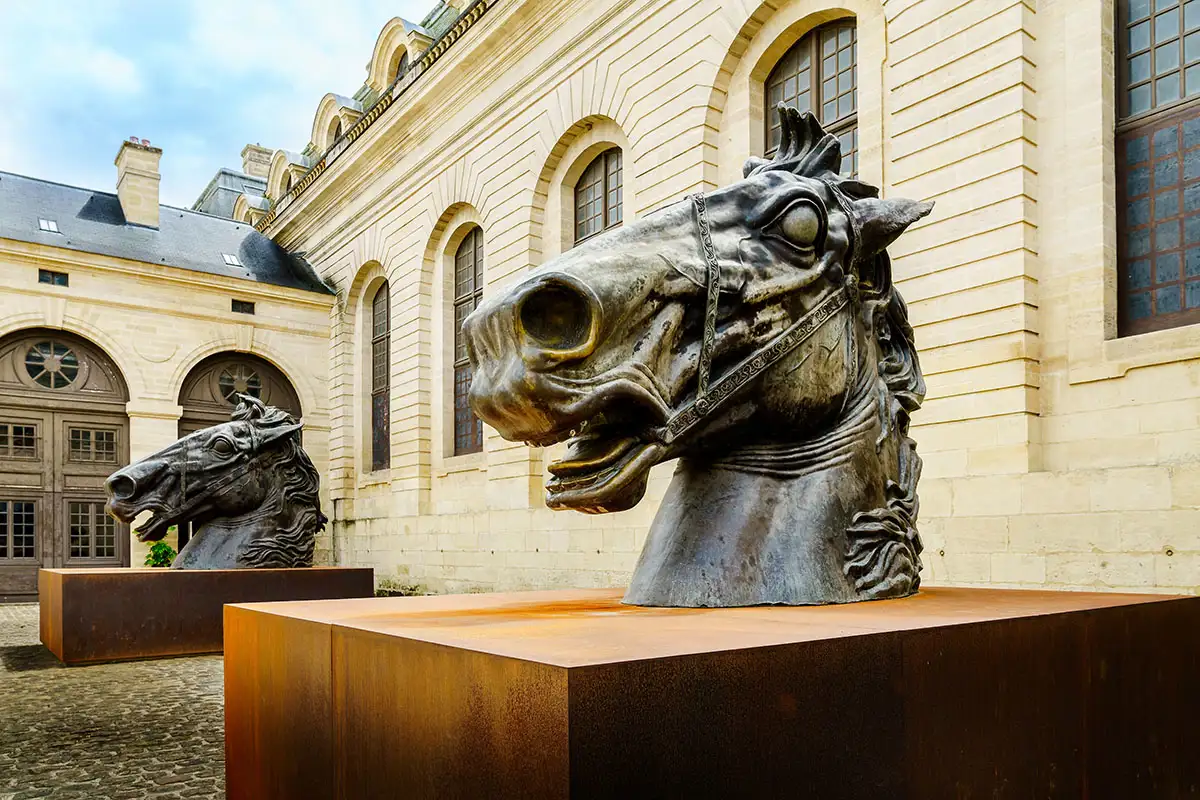 Two brass statues of horse heads on wooden plinths in front of a sandy coloured stone building