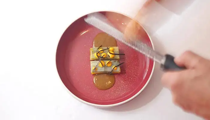 Hand verkruimelt brood boven een roze bord met twee kleine beige bietenbeignets met bruine saus 