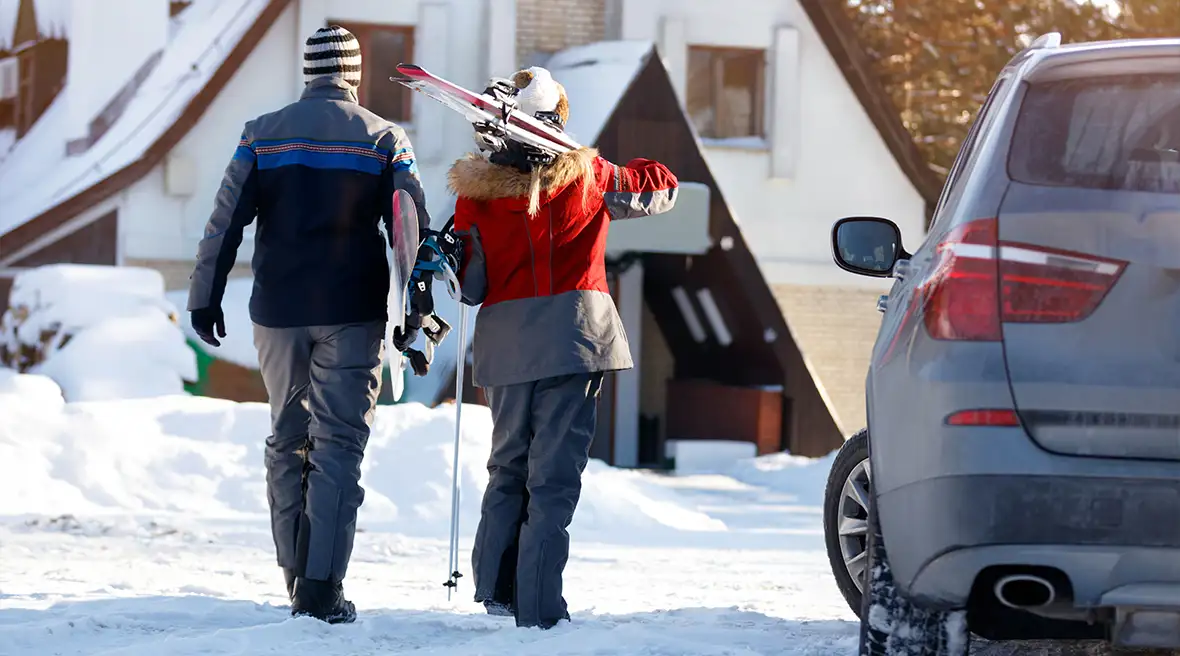 couple in winter wear walking to ski lodge with skis on shoulder car in foreground