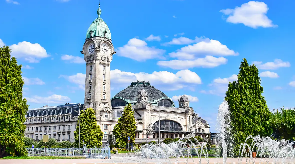 An impressive station building with a tall clock tower behind a public park with a water fountain