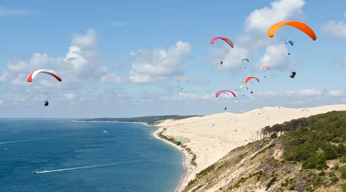 Numerous colourful paragliders sailing out over a huge sand dune and blue waters on a backdrop of blue sky with fluffy white clouds