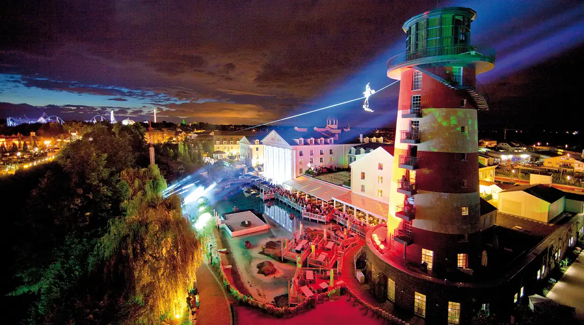 A lighthouse is illuminated at night above a hotel and swimming pool