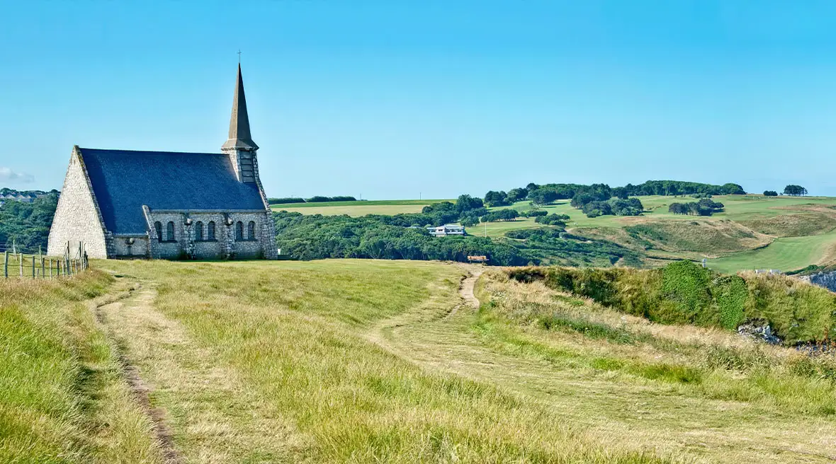 Panorama of Notre Dame de la Garde chapel and the cliff of Etretat, Normandy, France beneath a clear blue sky and surrounded by calm blue waters and grass