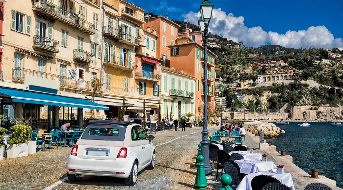 A white Fiat500 driving along the waterfront of a picturesque French town in the sun