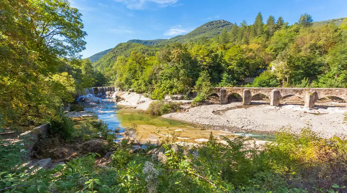 stone bridge on a pebbled beach next to a river surrounded by trees and hills under a sunny sky