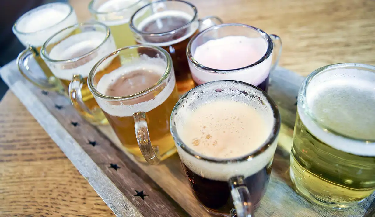 A wooden tray on a table with eight different glasses of beer on it in shades of yellow and brown