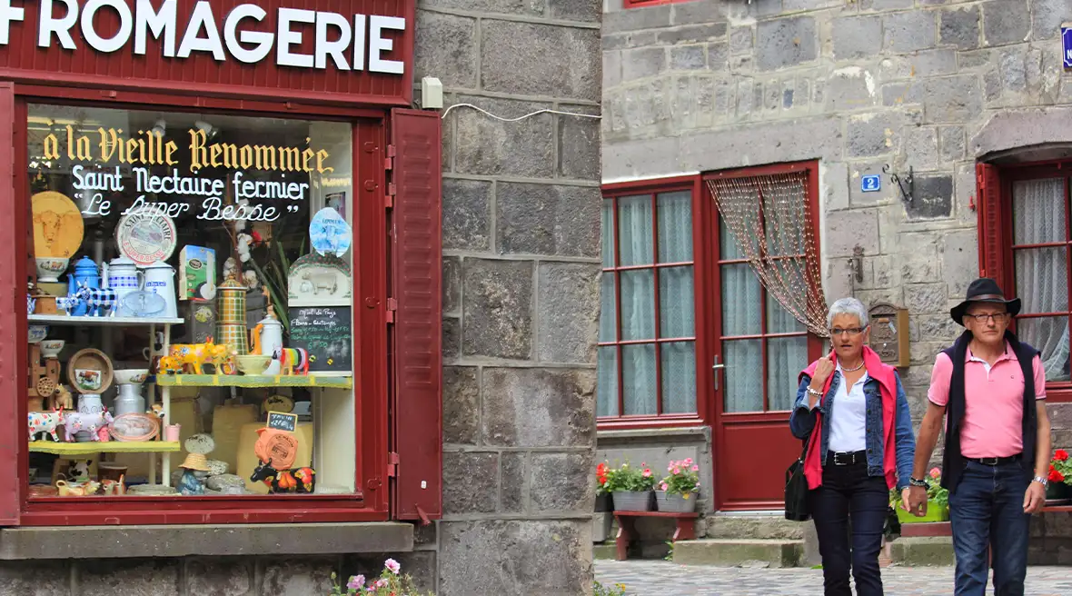 Two people walk down a street towards a cheese shop in a grey-brick building with dark red paintwork