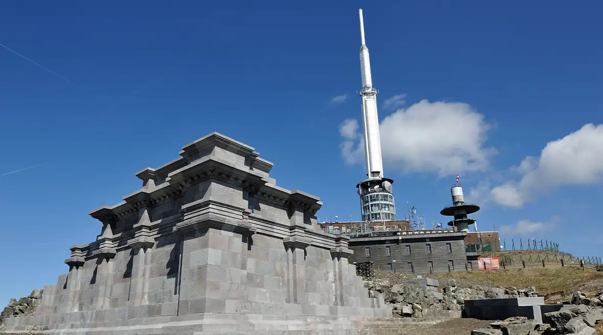 Grey stone structure with a large white spire behind it against a blue sky on top of a volcano