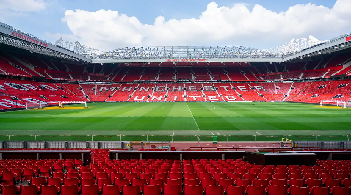 Binnenkant van stadion Old Trafford, met rode stoelen op de voorgrond, een grasveld en in enorme witte letters de tekst Manchester United