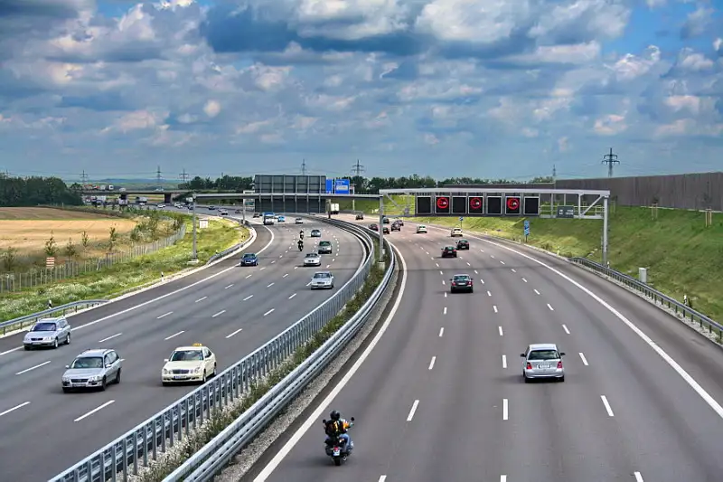 The A9 highway in Germany with four lanes of traffic moving in each direction. Greenery surrounds the roads and signs are visible in the distance. Vehicle are travelling along the road.