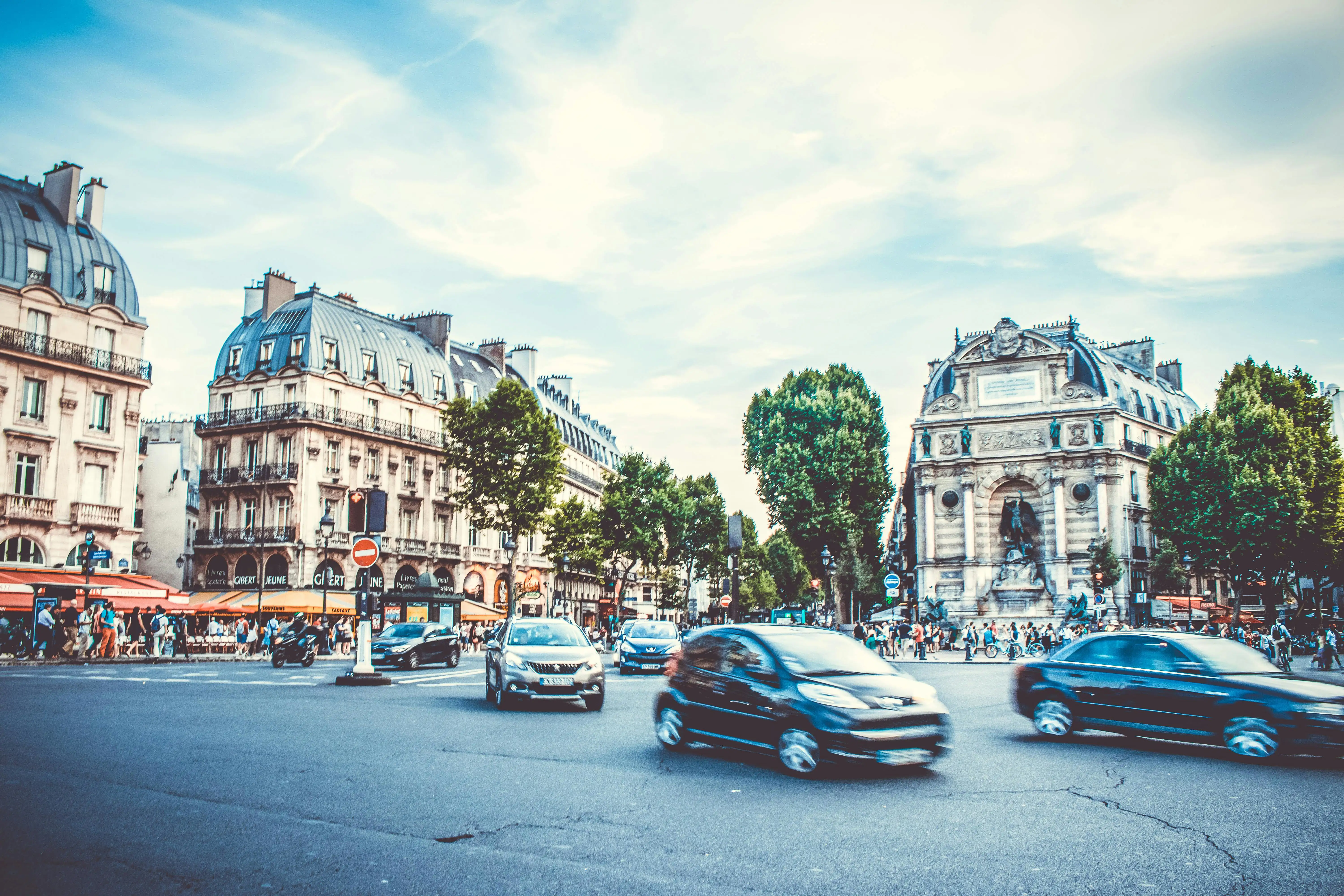 Cars driving through an intersection in Paris. Busy streets and the Fontaine Saint-Michel fountain is in the background.